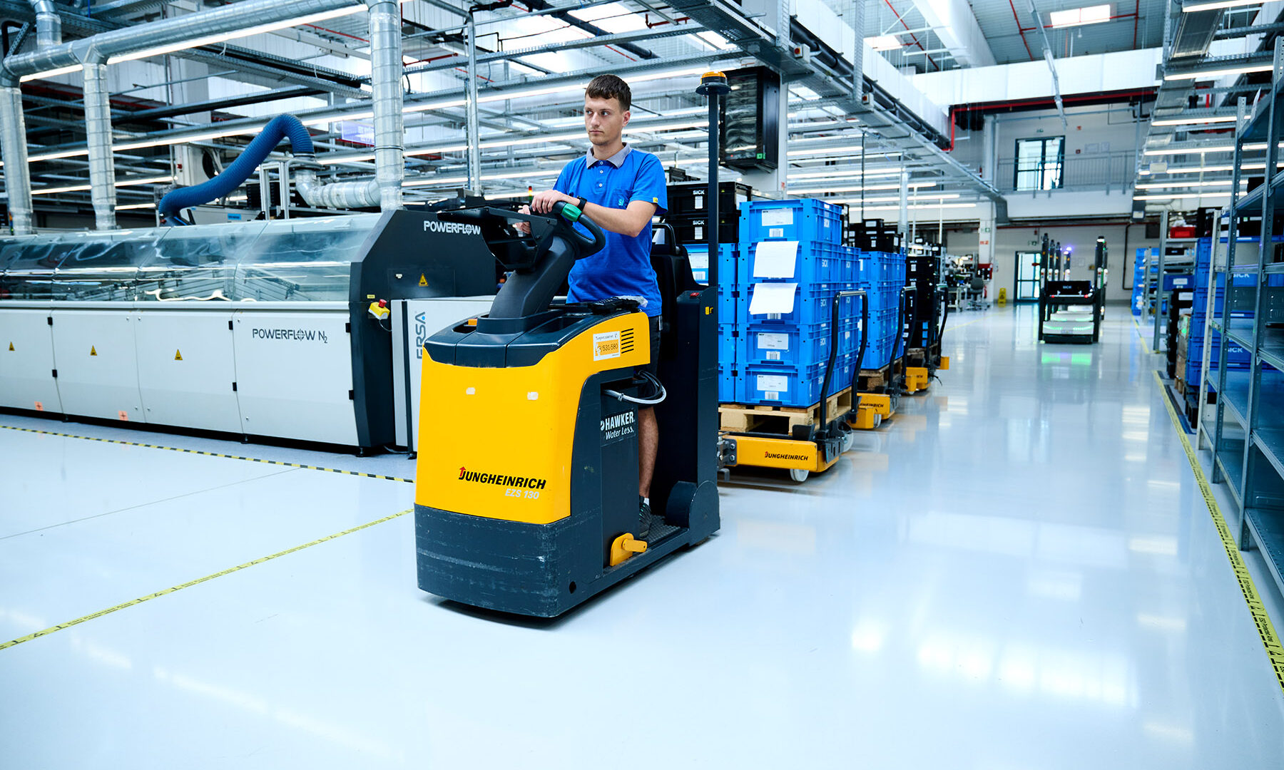 Un homme en chemise bleue conduit un tracteur électrique Jungheinrich jaune, tirant des bacs bleus sur des chariots à travers une installation industrielle moderne et lumineuse avec des machines et un stockage organisé.