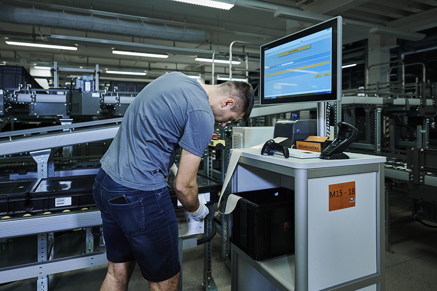 Retrieval of goods from the Evo Shuttle at B&R A man is standing at a work station with a labeling machine and monitor next to a conveyor with containers.