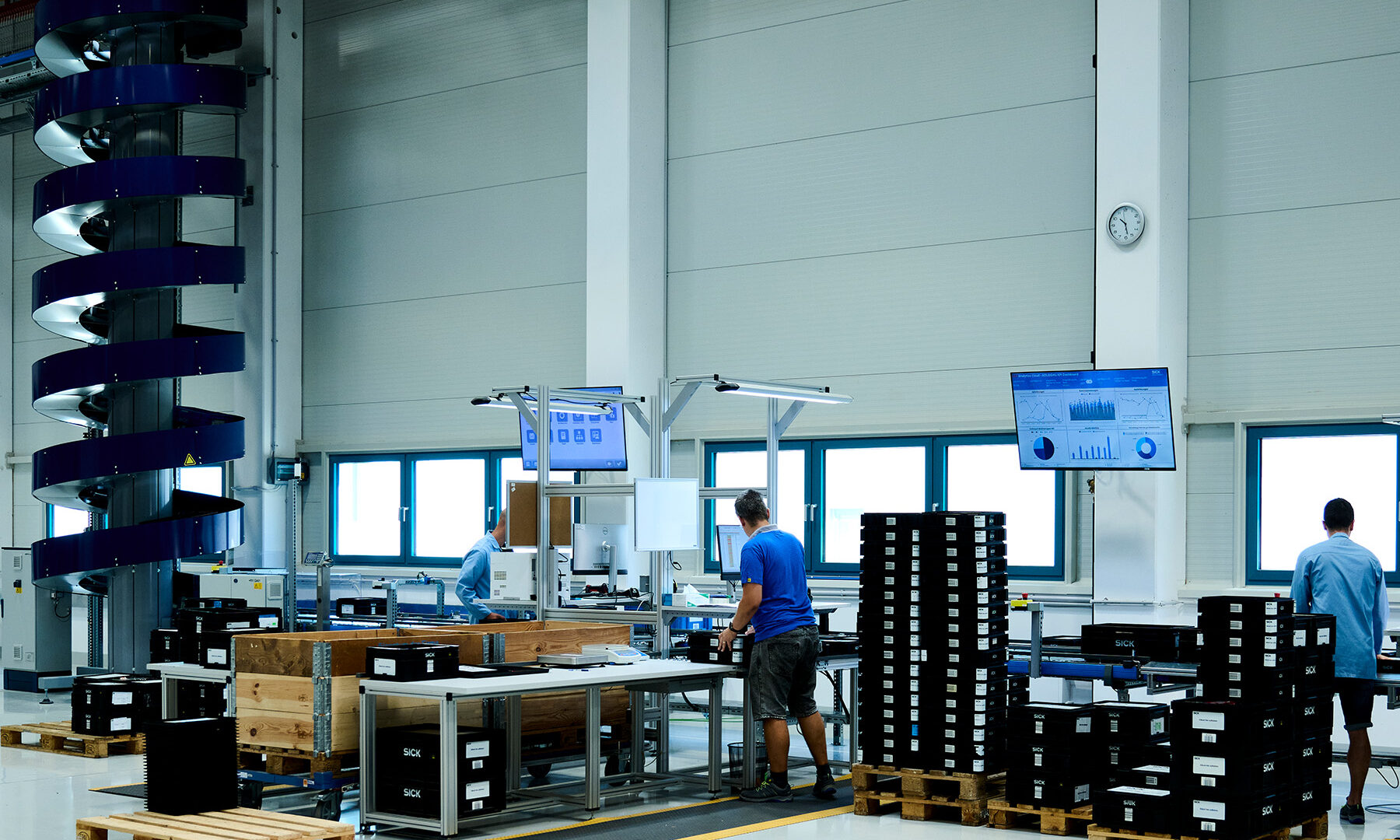 Un entrepôt moderne où des travailleurs manipulent des boîtes noires à des postes de travail, entourés de palettes et de caisses. Des écrans numériques affichent des données et un convoyeur bleu en spirale est visible sur la gauche. De grandes fenêtres et une horloge se trouvent sur le mur du fond.
