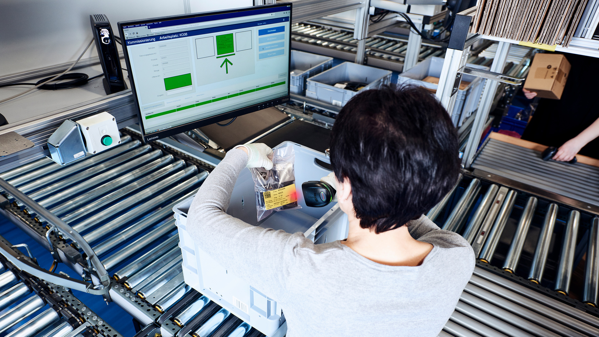Eine Person scannt ein Produkt an einem Arbeitsplatz mit einem Computermonitor, auf dem die Inventarsoftware von Retail Logistik angezeigt wird, umgeben von Förderbändern und Lagerbehältern in einem Lager oder Fulfillment Center.
