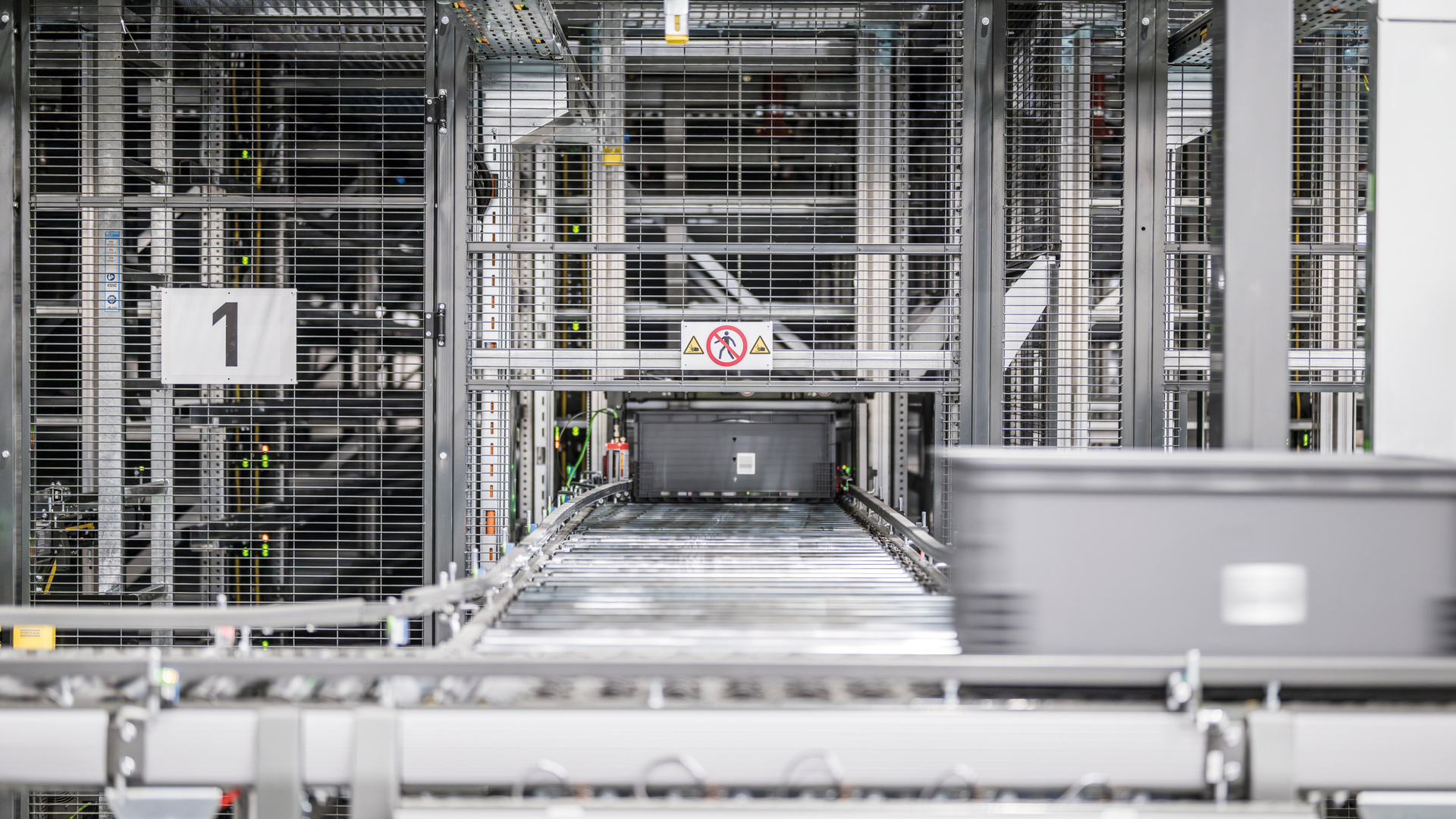 Automatisiertes Lager mit Metallregalen, Förderbändern und Lagerbehältern. Ein "Zutritt verboten"-Schild ist über der Förderstrecke zu sehen. Der Bereich wirkt sauber, modern und hochindustrialisiert.