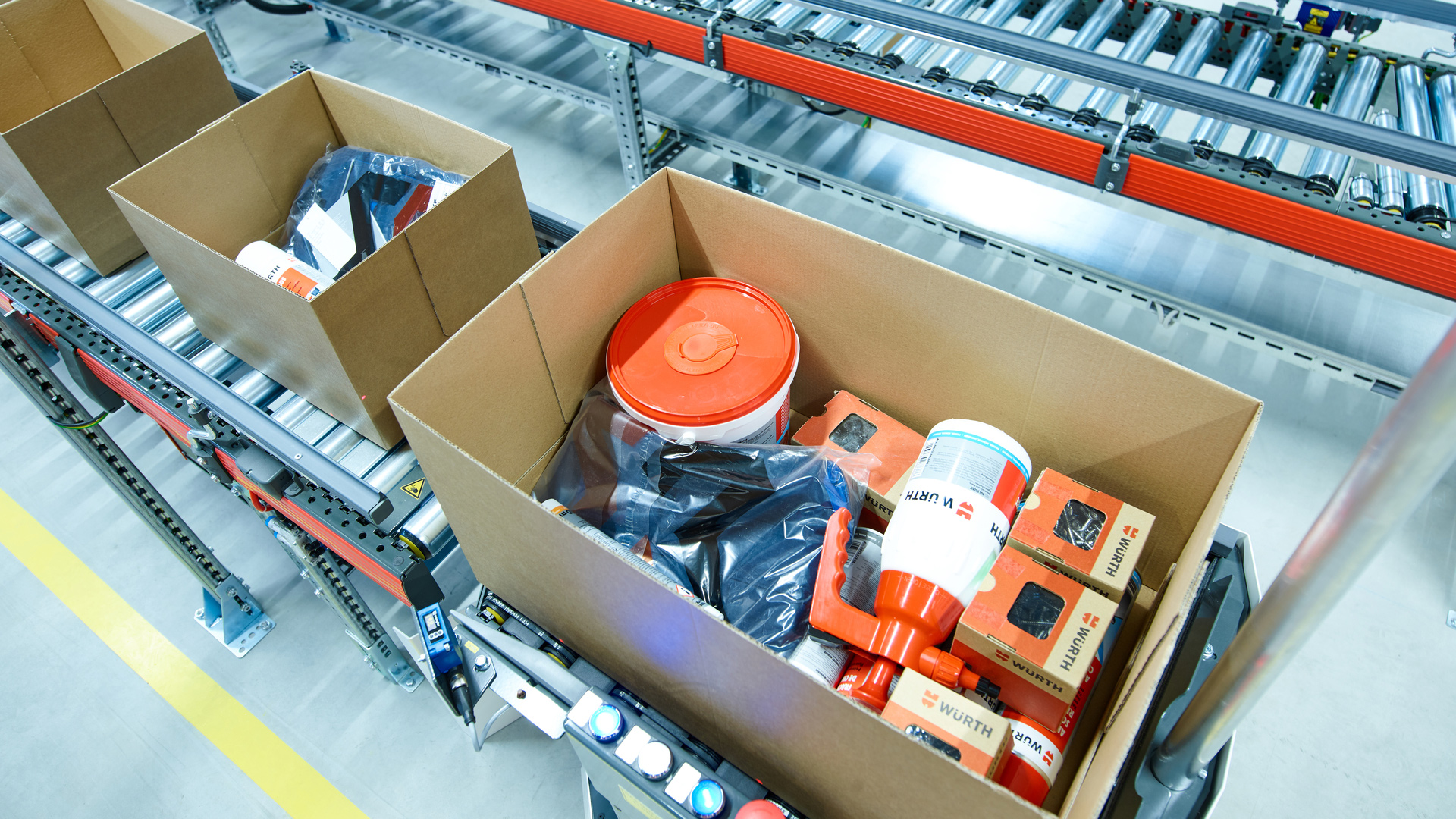 Offene Kartons auf einem Förderband in einem Lagerhaus, die verschiedene Produkte enthalten, darunter rote und weiße Behälter, schwarze Säcke und orangefarbene Verpackungen.