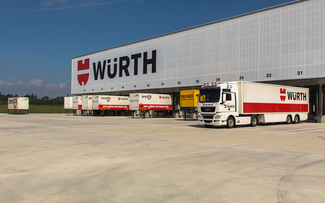 Several Würth trucks are parked at loading docks outside a large warehouse with a prominent Würth logo on the building. One Würth truck is driving past in the foreground on a sunny day.